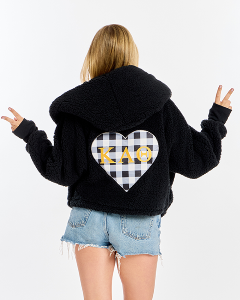 A model shown from the back wearing a black sherpa hoodie with a large heart-shaped black and white gingham pattern on the back, featuring the Greek letters 'Kappa Alpha Theta' in yellow. The model is also wearing light blue denim shorts against a plain white background.
