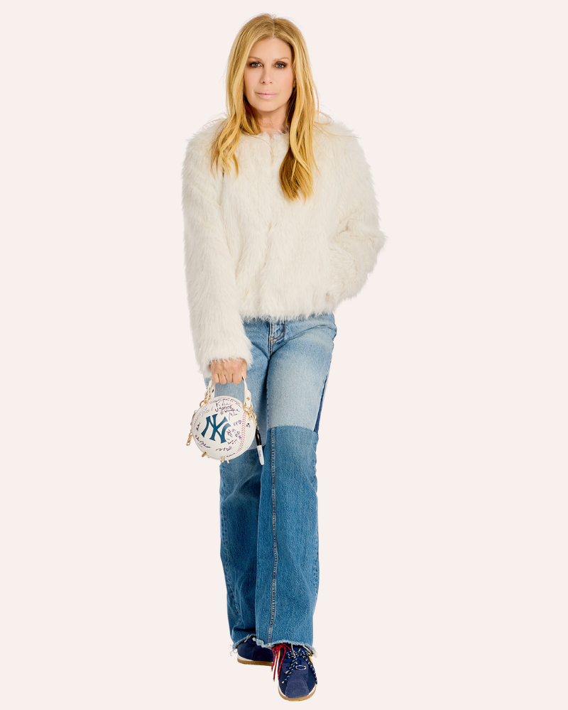 A woman stands against a plain light background wearing a fluffy white cropped sweater, blue jeans with a patchwork leg design, navy sneakers, and holding a small white handbag with a New York Yankees logo.