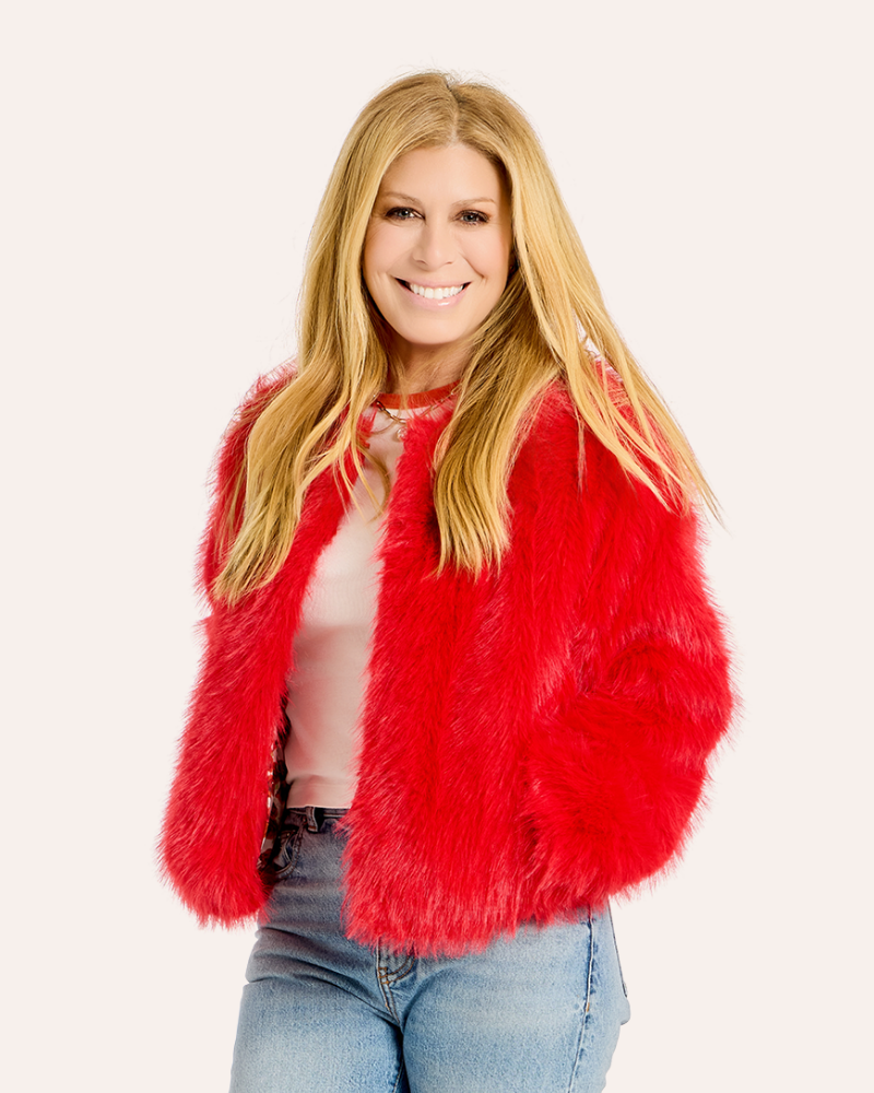 A smiling blonde woman wearing a bright red faux fur jacket over a light pink top and blue jeans, photographed against a plain light background.