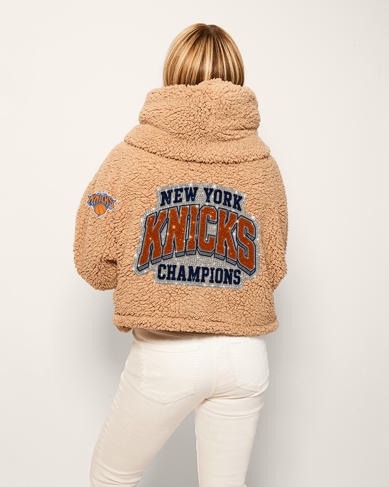 Person wearing a tan sherpa jacket with 'New York Knicks Champions' text on the back against a white background