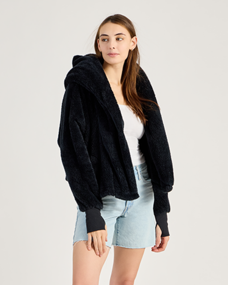 Woman wearing a black hoodie over a white top and light blue shorts on a plain background