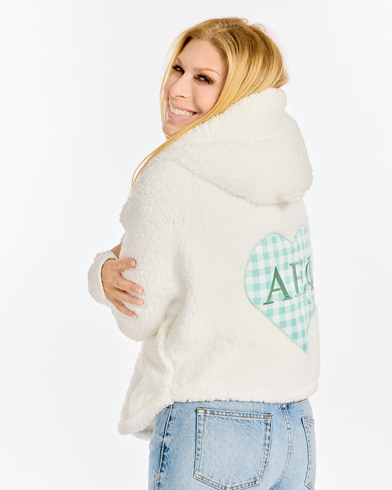 A woman wearing a white sherpa fleece hoodie with a green and white gingham heart graphic on the back, paired with light blue jeans, photographed against a solid white studio background.