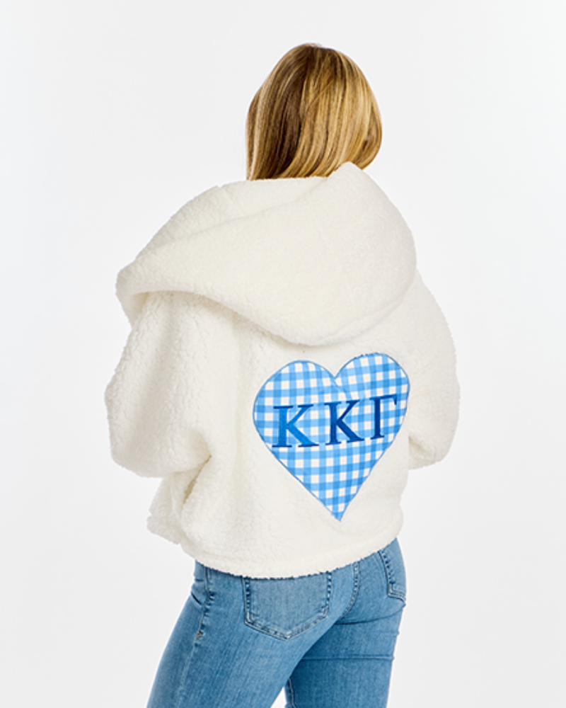 A woman viewed from behind wearing a cream sherpa hoodie with a blue and white gingham heart on the back featuring the letters 'Kappa Kappa Gamma' in blue, paired with blue jeans against a white studio background.