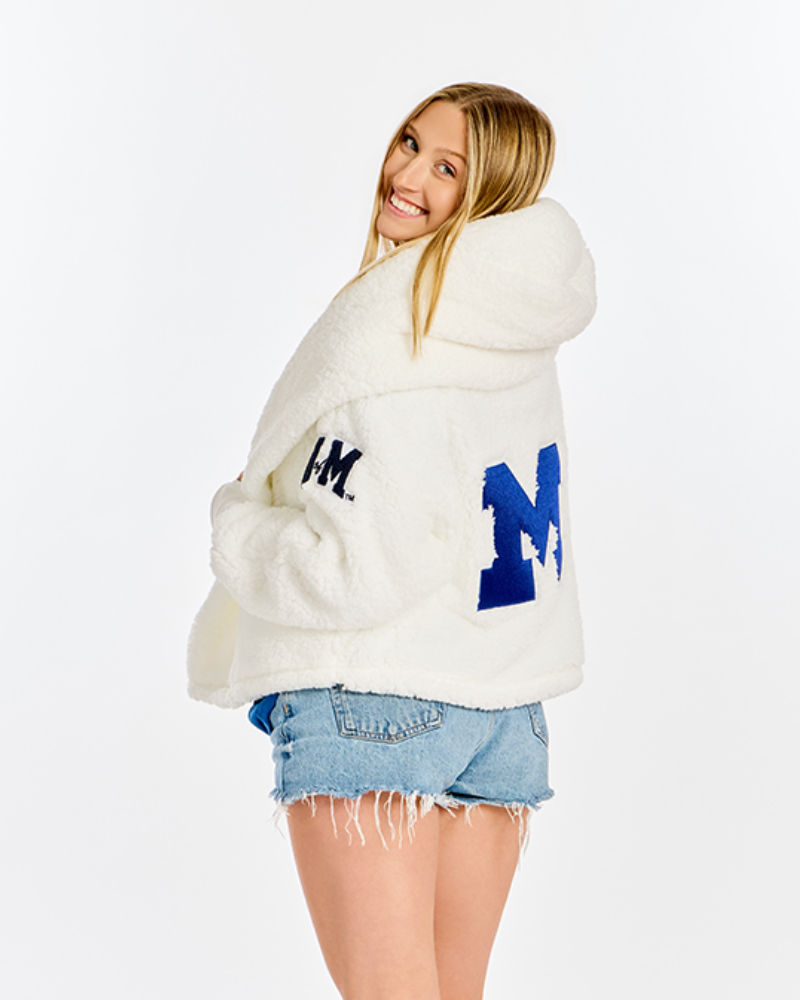 A woman modeling a white sherpa fleece hooded cardigan with blue collegiate letter 'M' embroidery on the back and a small navy 'U of M' logo on the bicep, paired with denim shorts against a white studio background.