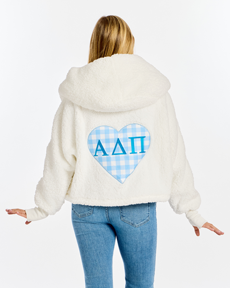 A woman shown from the back wearing a cream sherpa hoodie with a blue gingham heart patch on the back featuring the Greek letters Alpha Delta Pi, paired with blue jeans against a white studio background.