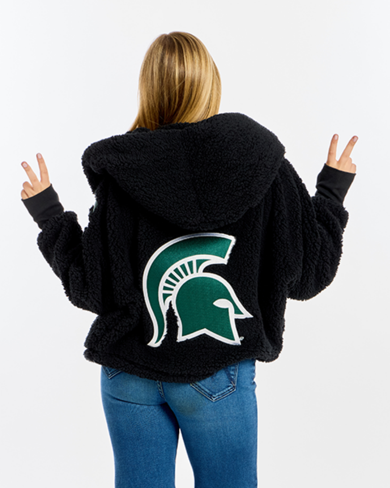 A woman shown from behind wearing a black sherpa fleece hoodie with a large green and white Michigan State Spartans helmet logo on the back, paired with blue jeans against a white studio background.