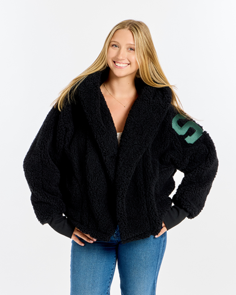 A smiling blonde woman wearing a black sherpa lounger jacket with a green 'S' on the sleeve, paired with blue jeans, against a plain white studio background.