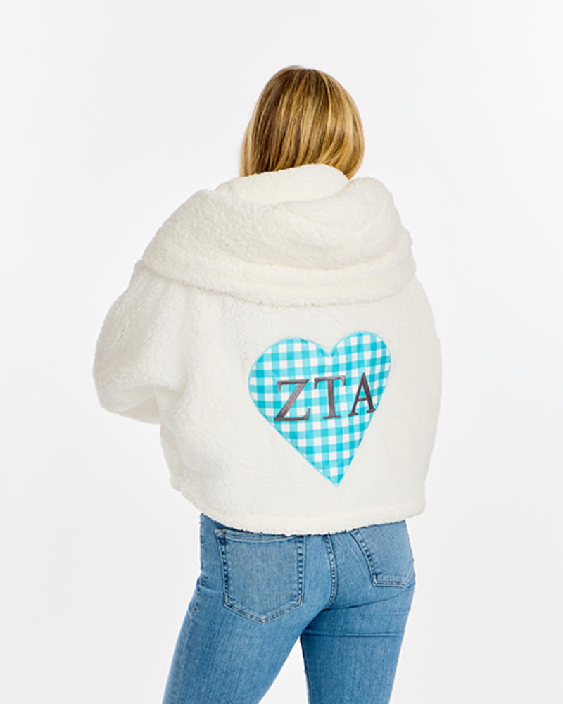 A woman viewed from behind wearing a cream sherpa hoodie with a blue and white gingham heart graphic on the back featuring the letters 'ZTA' in brown, paired with blue jeans, against a plain white background.