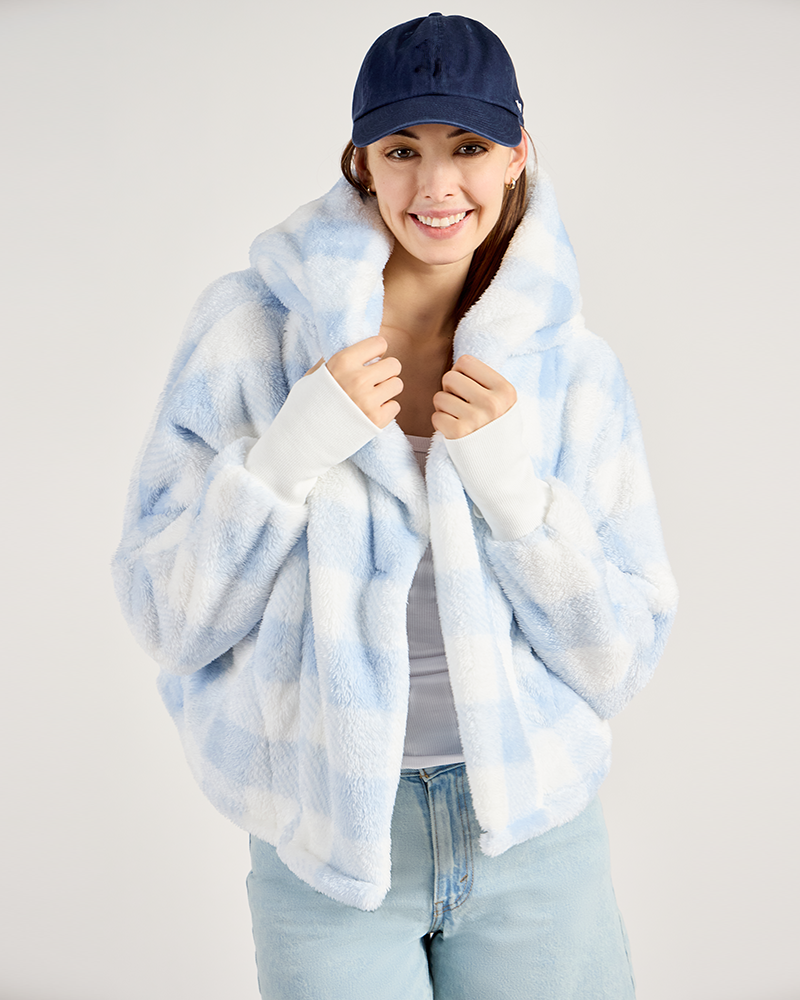 Woman wearing a light blue and white gingham jacket over a white shirt, with a navy cap on a plain background.