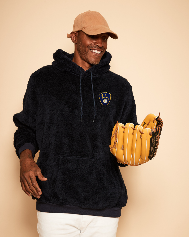 Man wearing a navy hoodie with a milwaukee brewers logo, beige cap, and holding a baseball glove against a beige background