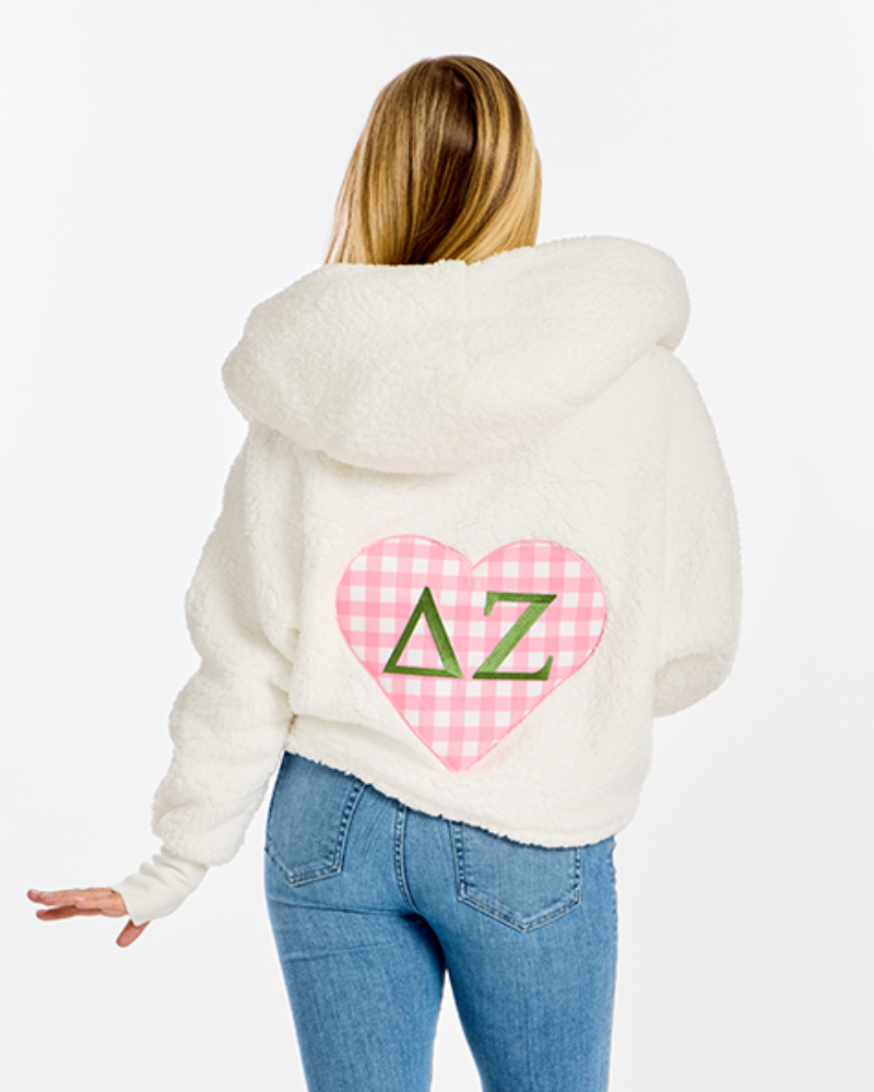 A woman viewed from behind wearing a cream-colored fuzzy hooded sweatshirt with a pink gingham heart patch on the back featuring green Greek letters Delta Zeta, paired with blue jeans against a white studio background.