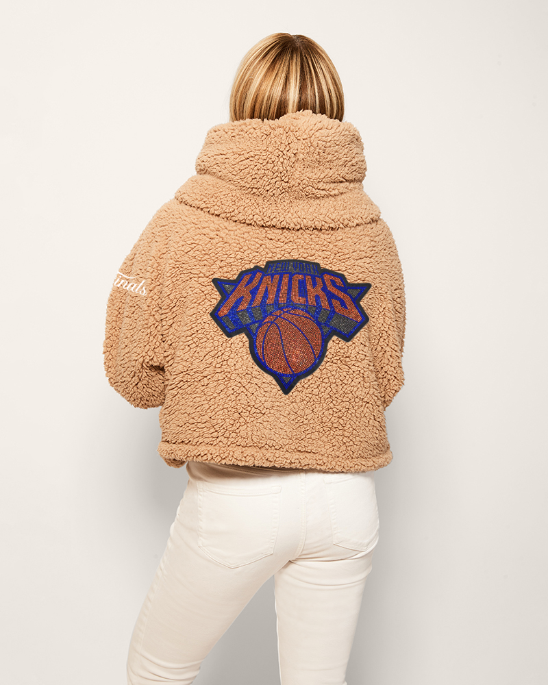 Person wearing a beige sherpa jacket with a Knicks logo on the back against a white background