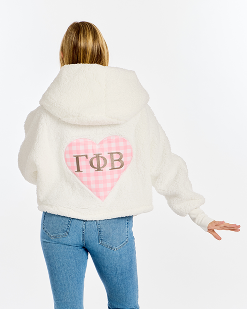 A woman shown from the back wearing a cream sherpa hoodie with a pink gingham heart and Greek letters 'Gamma Phi Beta' on the back, paired with blue jeans, against a plain white background.