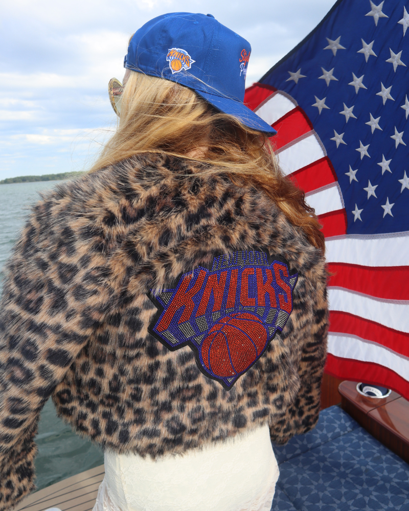 Person wearing a leopard print jacket with a Knicks logo, standing next to an American flag by water.