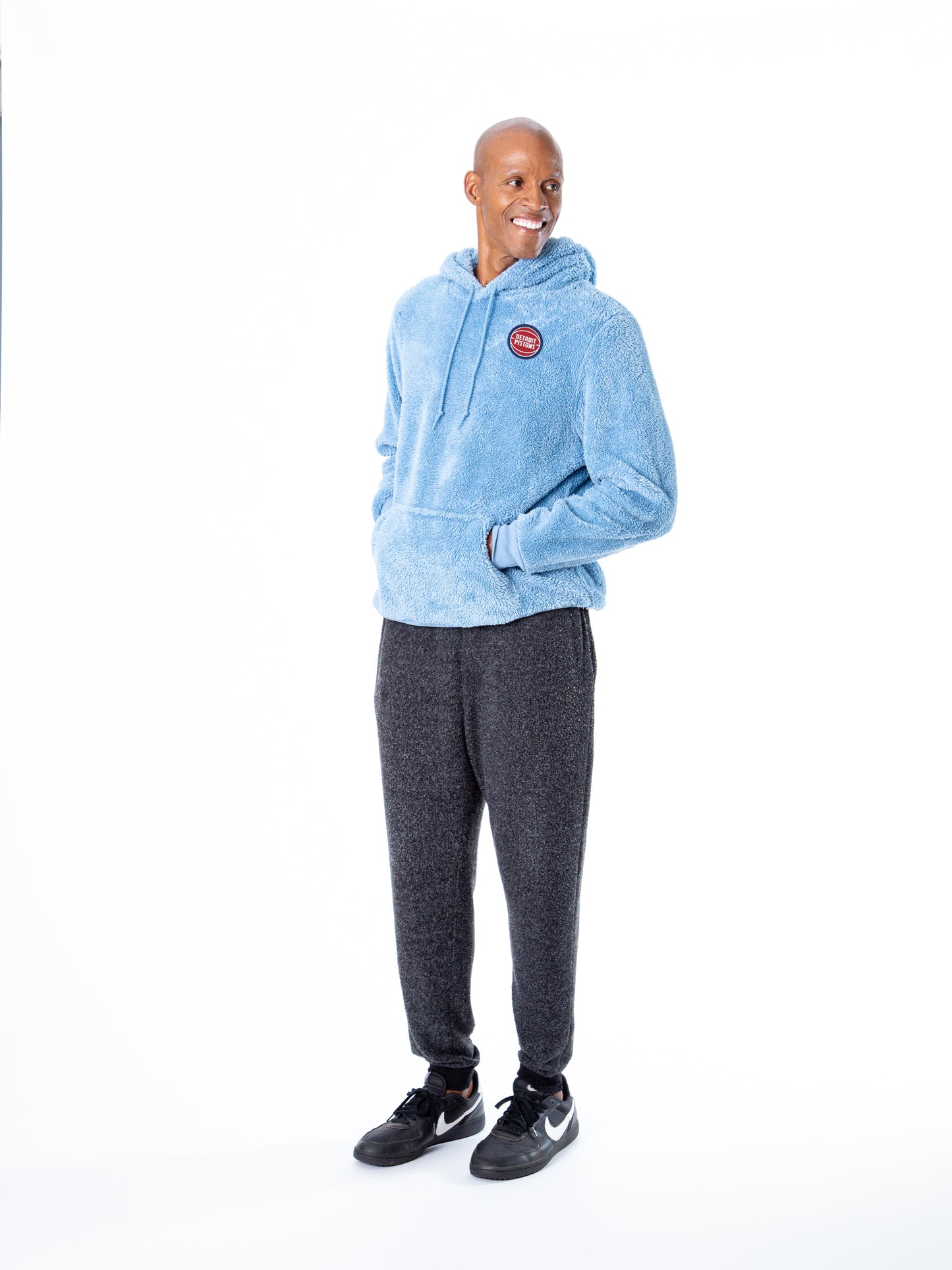 Side view of man with hands in the hoodie pocket wearing light blue sherpa hoodie with a front kangaroo pocket and drawstring hood, featuring small circular Detroit Pistons embroidery on the left chest.