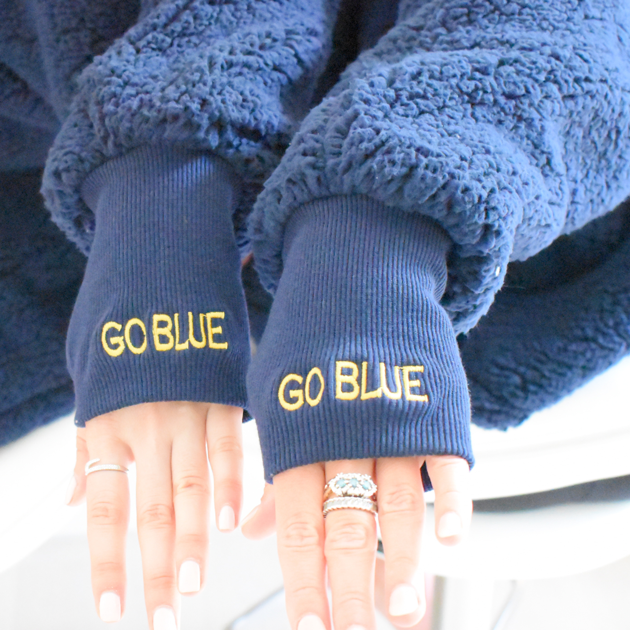 Close-up of two hands wearing a navy blue sherpa lounger with ribbed cuffs, both embroidered with the phrase "GO BLUE" in yellow thread. The person is seated, with white manicured nails and multiple rings on their fingers.