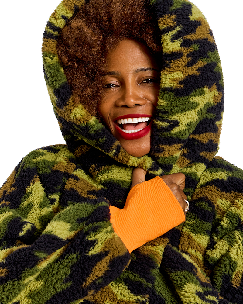 A smiling woman wearing a plush hooded jacket in a green, olive, and black camouflage pattern, holding up an orange sleeve to shield her face from the camera, against a solid white background.