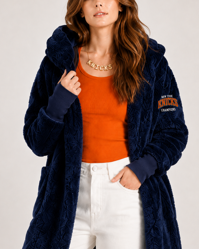 Woman wearing a navy robe with 'Knicks' branding over an orange top and white pants on a light gray background