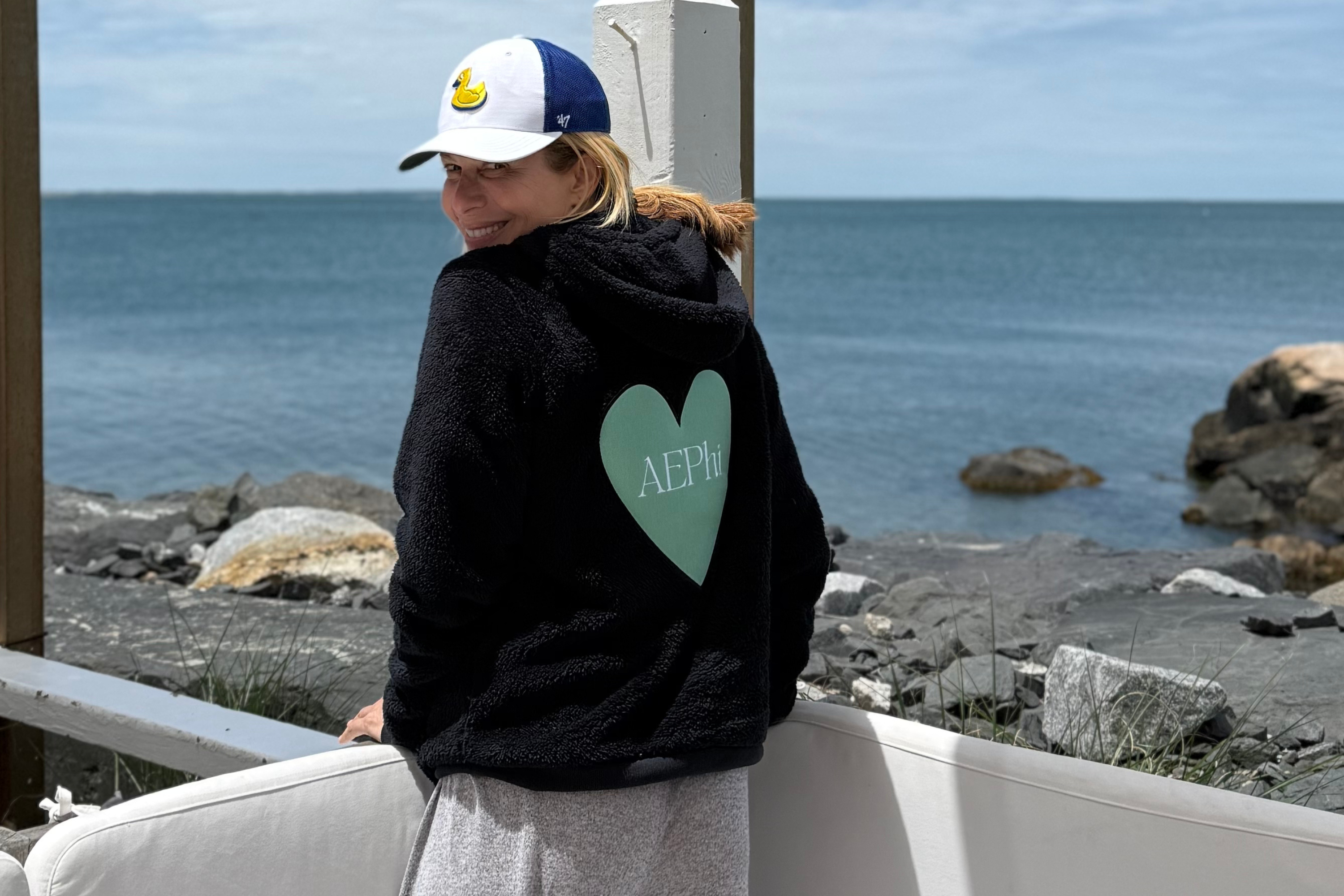 Person wearing a black hoodie with a green heart and 'AEPHi' text, sitting by the ocean.