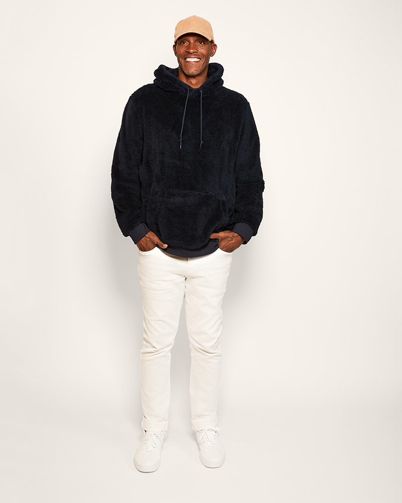 Person wearing a navy hoodie, white pants, and a beige cap on a plain background