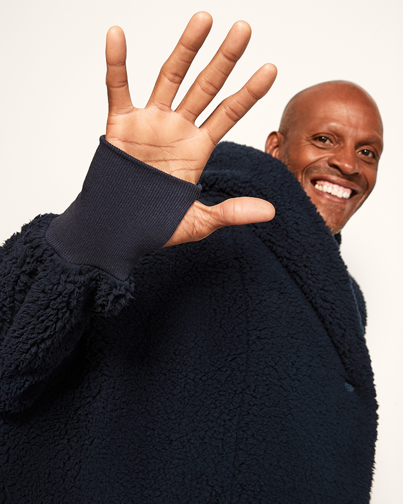 Man wearing a dark blue fleece jacket with his hand raised, showing five fingers against a plain background.