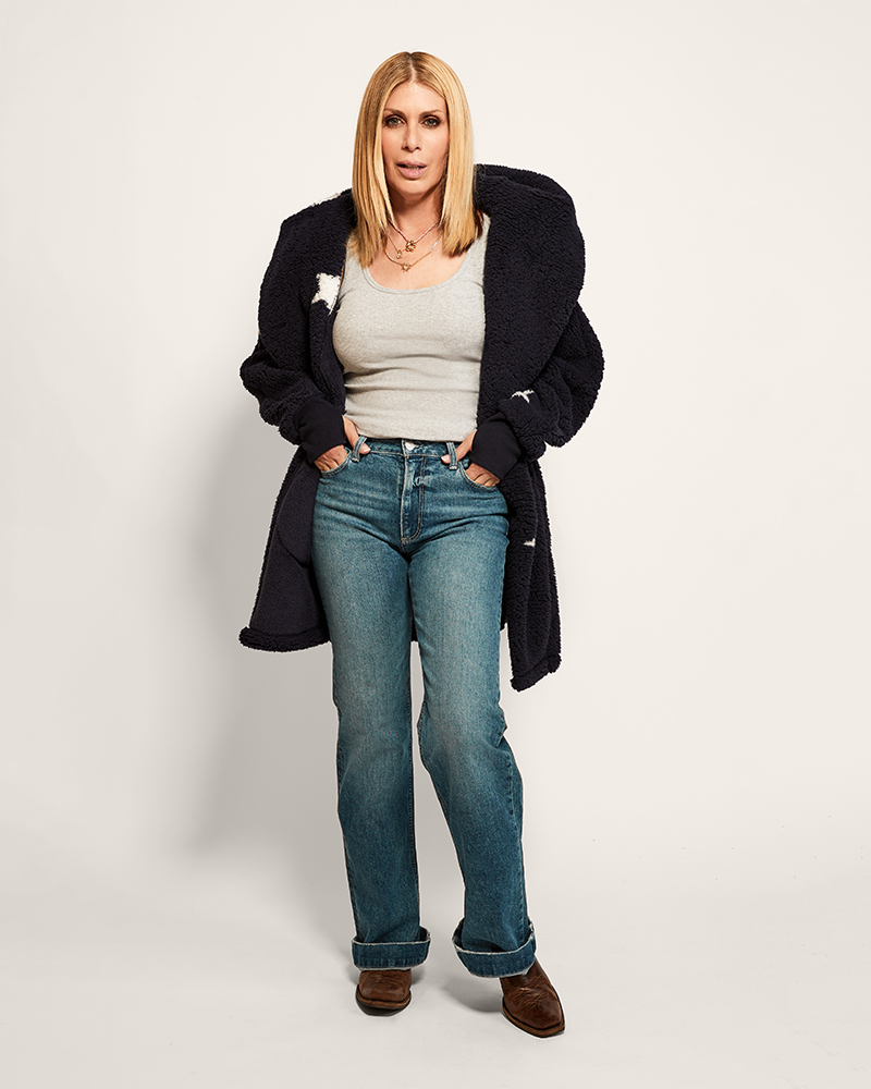 Woman wearing a navy coat with star pattern, white shirt, blue jeans, and brown shoes on a plain background