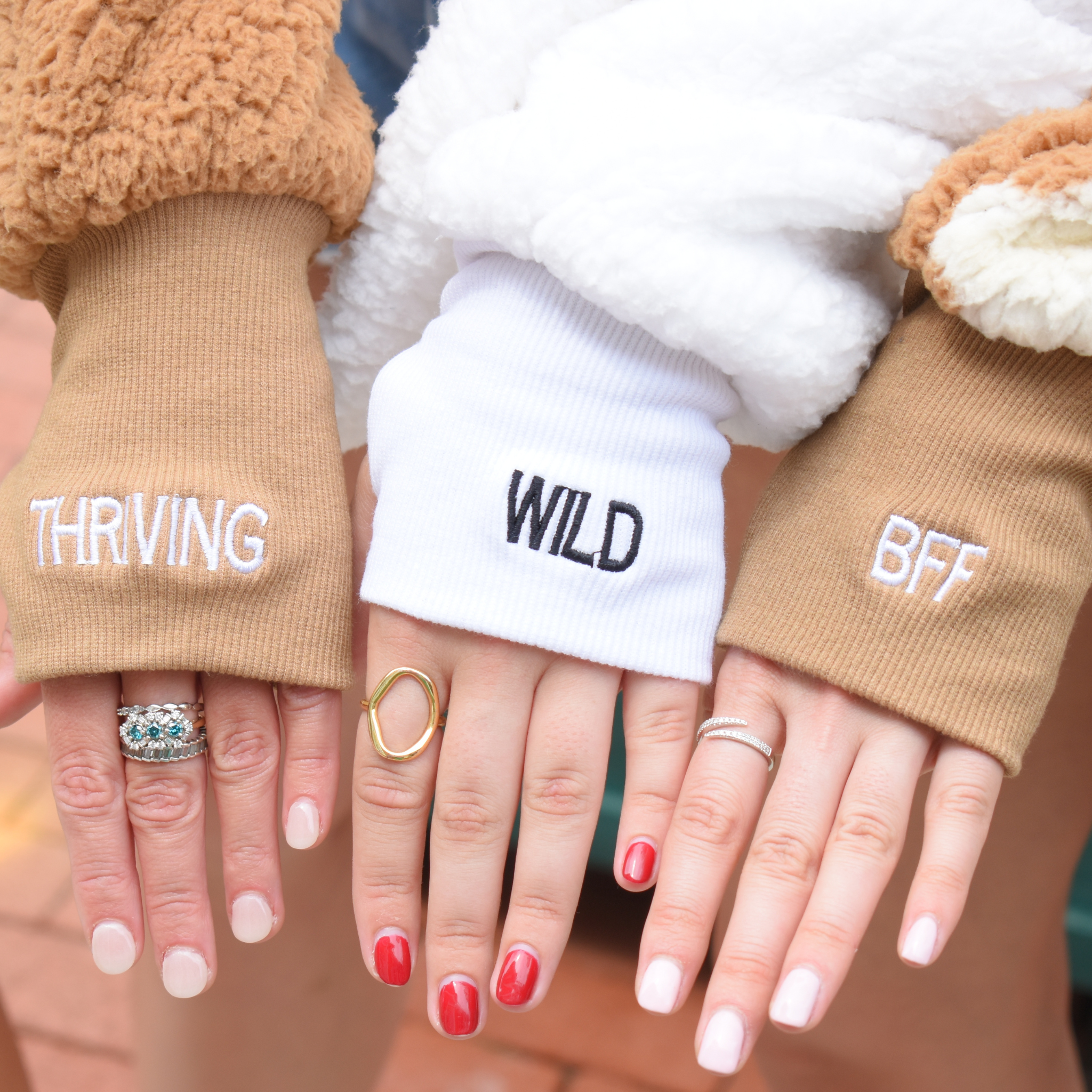 multiple hands grouped together showing off cuff embroidery. From left to right the cuff embroidery reads "thriving", "wild" and "bff"