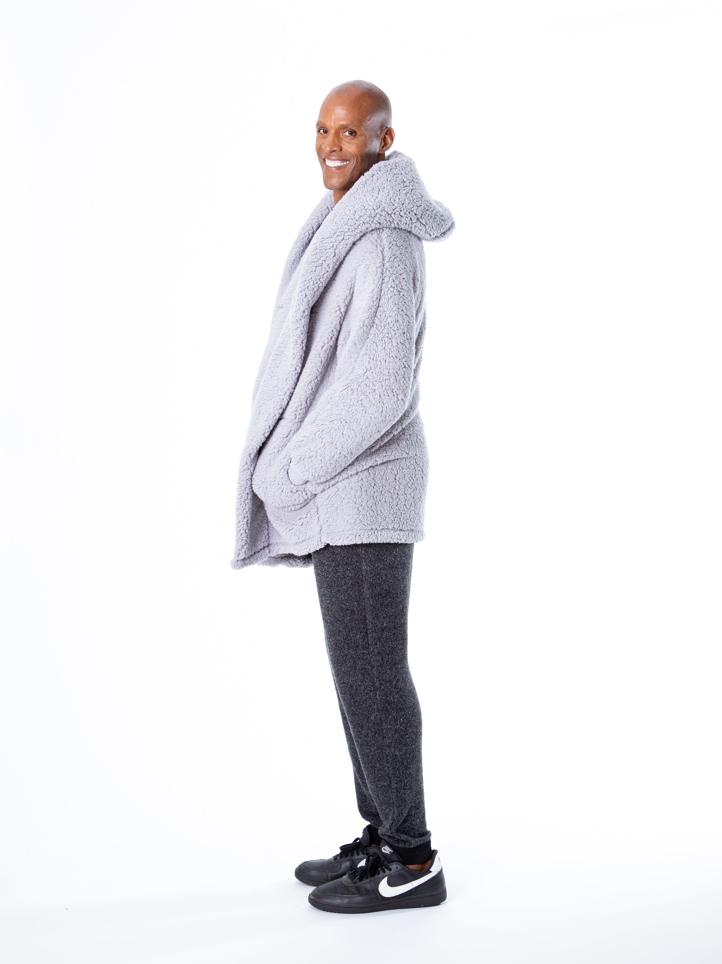 A person is standing sideways, smiling, wearing a cozy Chateau Grey Classic Lounger by Jill Martin, dark grey sweatpants, and black Nike sneakers against a plain white background.
