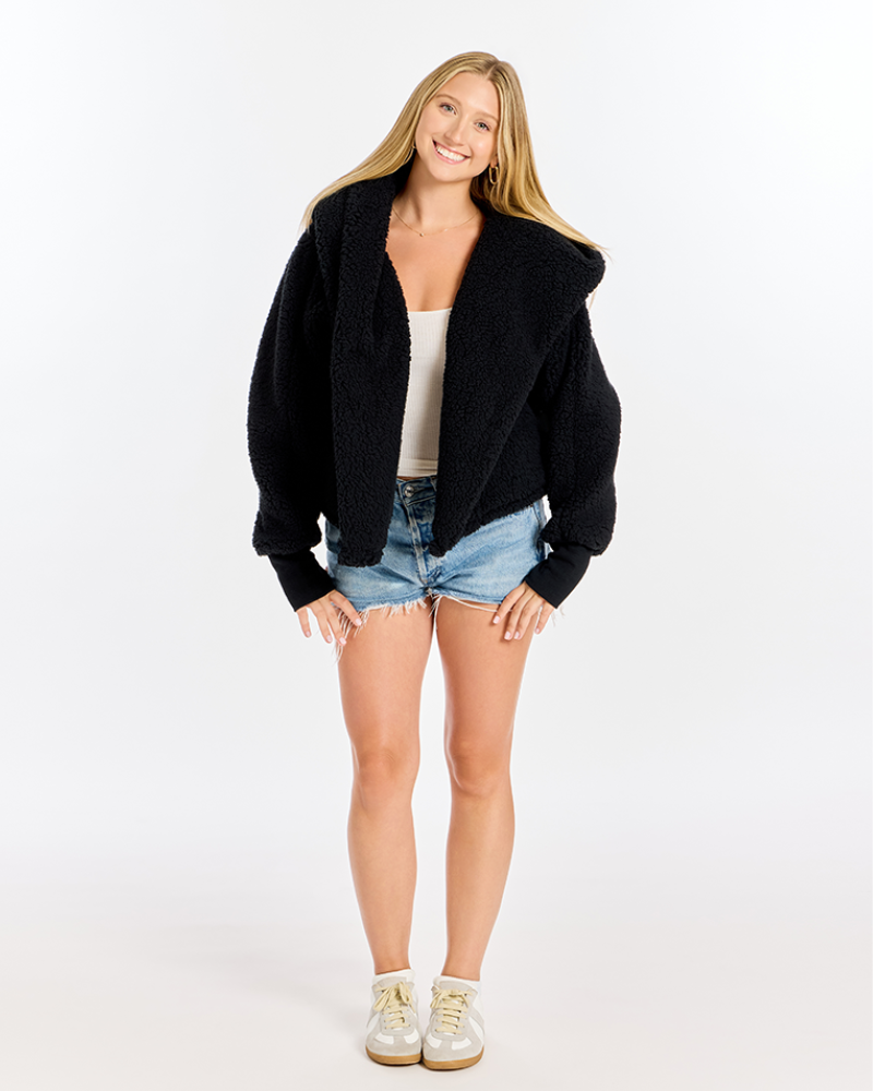 A woman modeling a black fuzzy cropped cardigan with a wide collar, worn open over a white tank top, denim shorts, and white sneakers, against a plain white studio background.