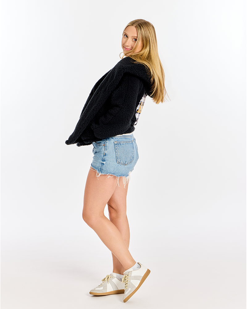 A woman posing in a black knit sweater, light blue denim shorts, and white sneakers against a plain white studio background.