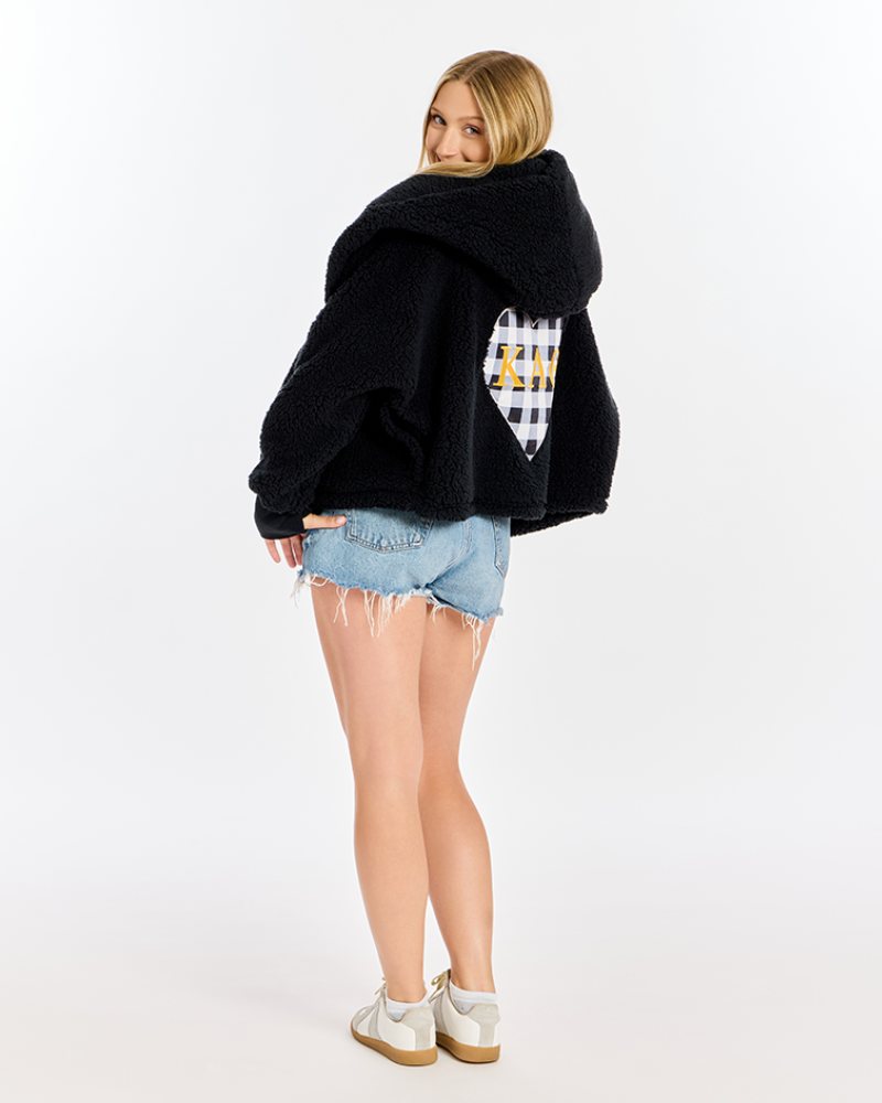 A woman modeling a black fuzzy cropped hoodie with a white and black checkered heart patch on the back, paired with denim shorts and white sneakers, against a plain white studio background.