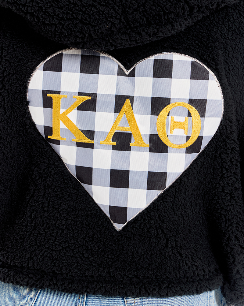 Close-up of a black sherpa hoodie featuring a large heart-shaped patch with a black and white gingham pattern and gold Greek letters 'Kappa Alpha Theta' embroidered across the front.