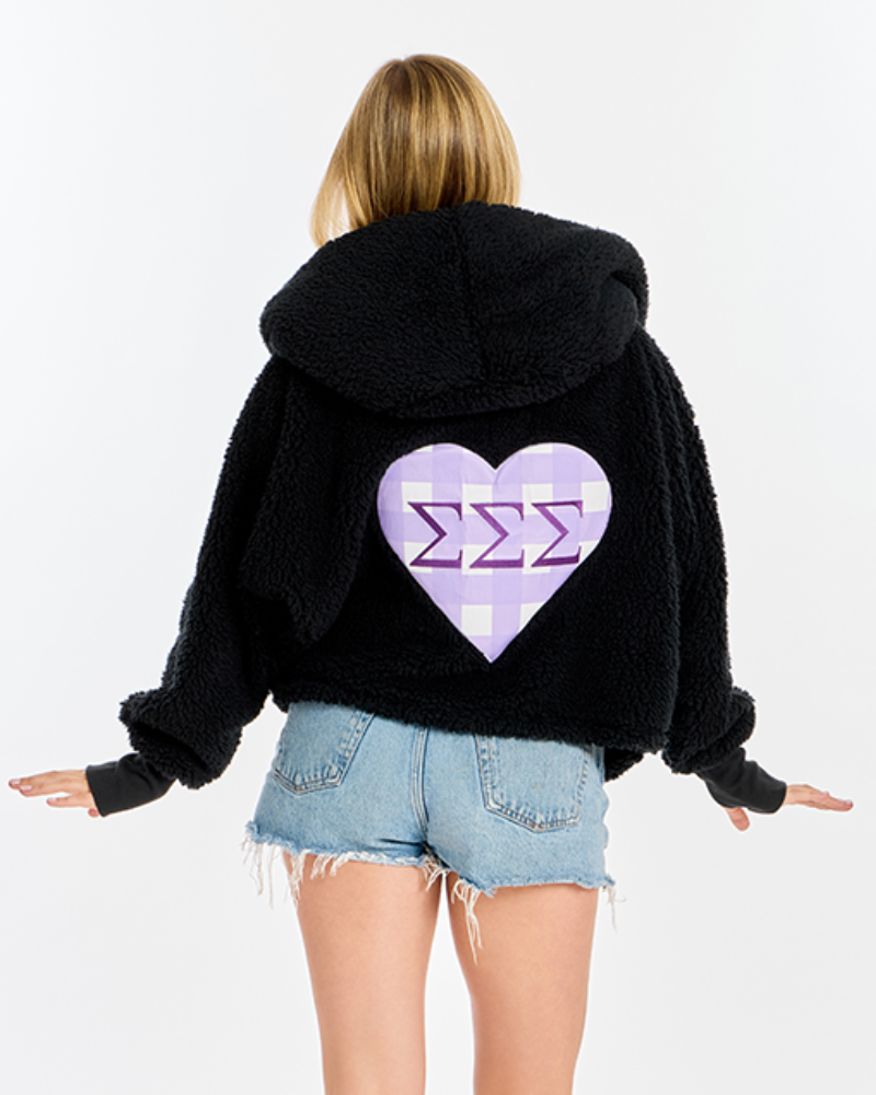 A woman shown from the back wearing a black fuzzy hooded sweatshirt with a large lavender and white heart graphic containing the Sigma Sigma Sigma Greek letter symbol, paired with denim shorts against a plain white background.