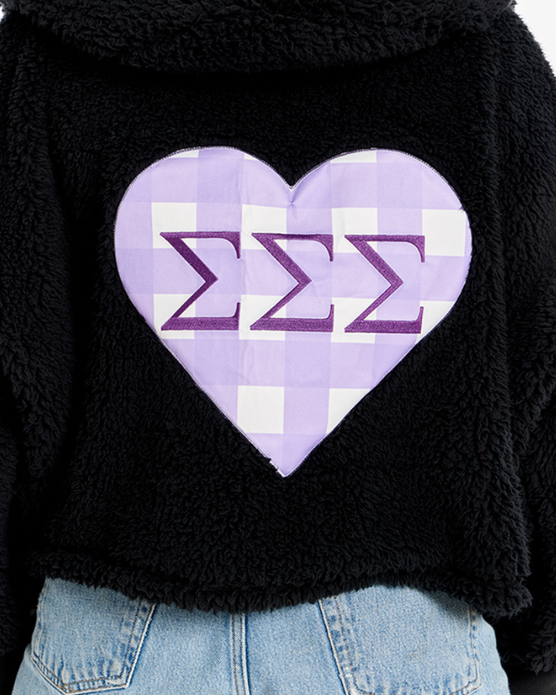 A close-up of a black fuzzy cropped hoodie featuring a large lavender and white checkered heart patch on the back with purple Greek letters Sigma Sigma Sigma.