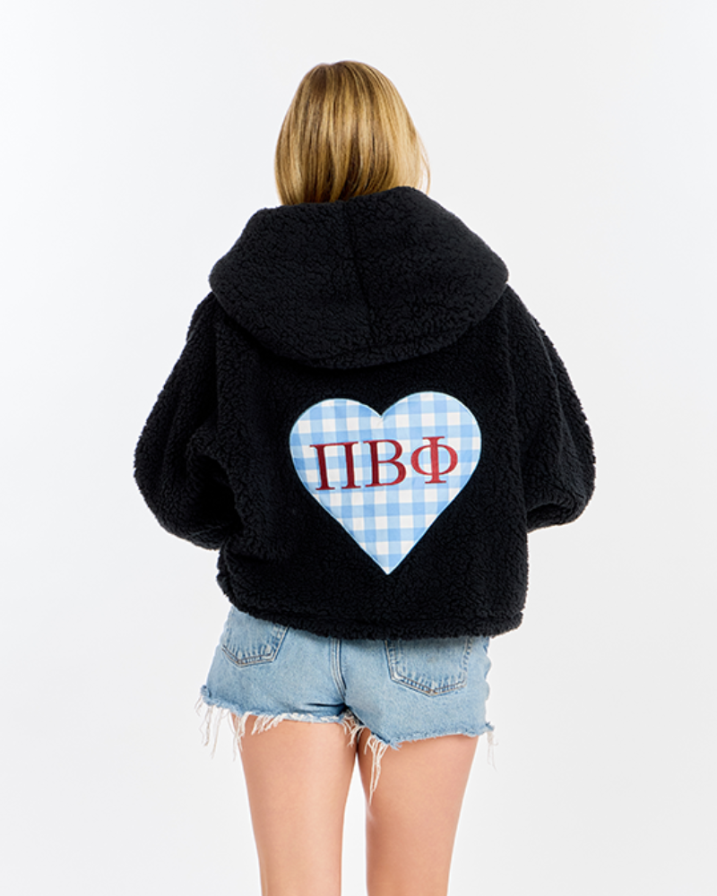 A woman viewed from behind wearing a black sherpa fleece hoodie with a large heart-shaped Greek letter graphic on the back, paired with denim shorts, against a plain white background.