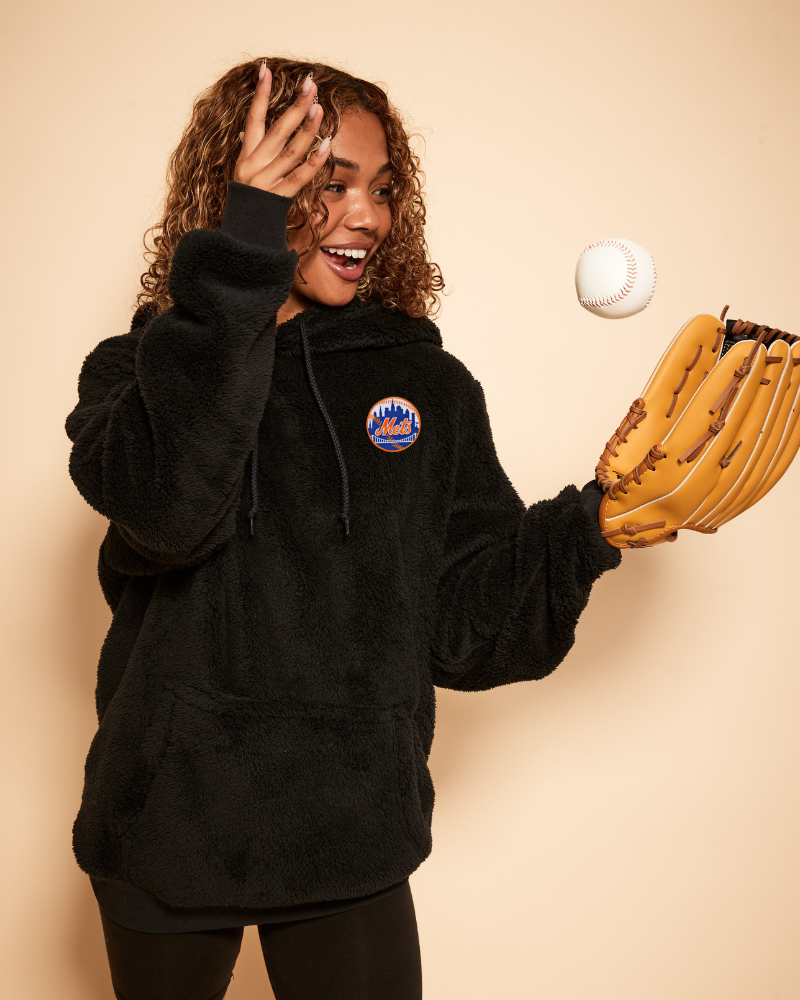 Person wearing a black hoodie with a new york mets logo, holding a baseball and a glove against a beige background