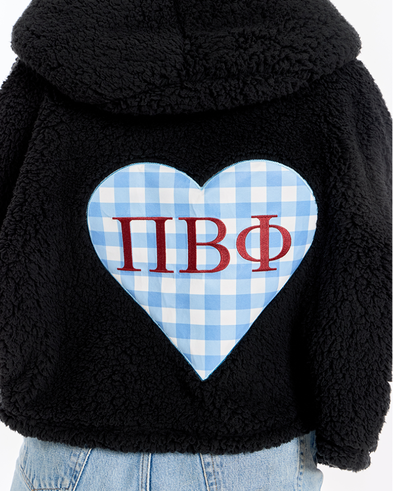 A close-up of a black sherpa fleece hoodie featuring a large light blue and white gingham heart on the back with the Greek letters 'Pi Beta Phi' in red, worn by a model with light blue jeans visible at the bottom.
