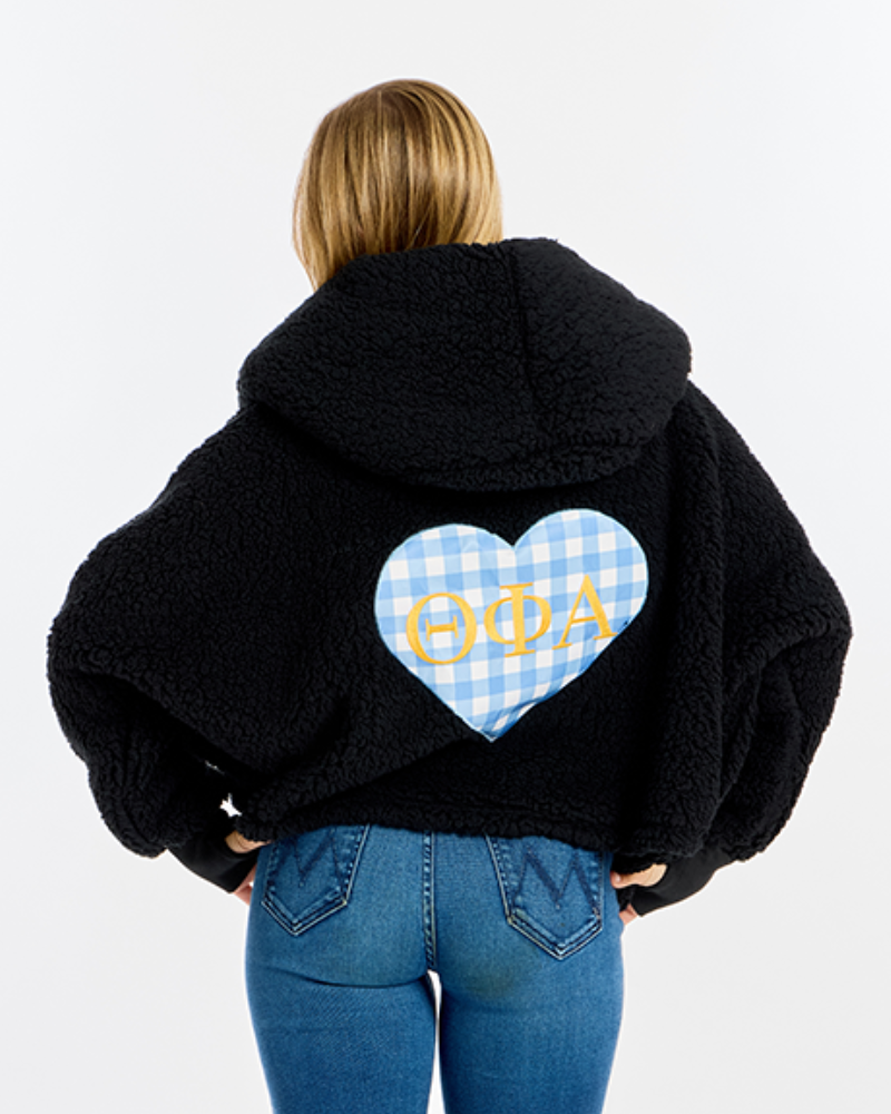 A woman viewed from behind wearing a black sherpa hoodie with a large light blue and white gingham heart on the back featuring gold Greek letters, paired with blue jeans, against a plain white background.