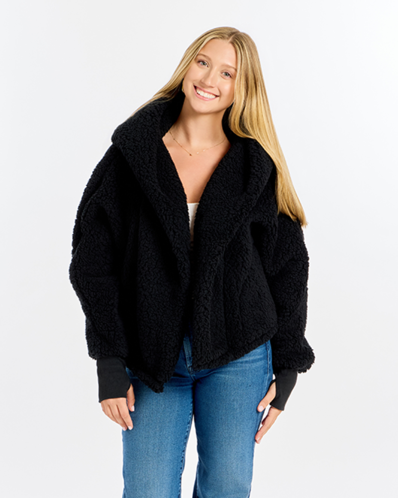 A smiling blonde woman wearing a black textured teddy fleece jacket with a large shawl collar, paired with blue jeans, against a plain white studio background.