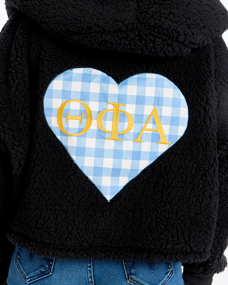 Close-up of a black sherpa hoodie featuring a large heart-shaped patch with a light blue and white gingham pattern and gold Greek letters 'Theta Phi Alpha' on the back, worn by a model in blue jeans.