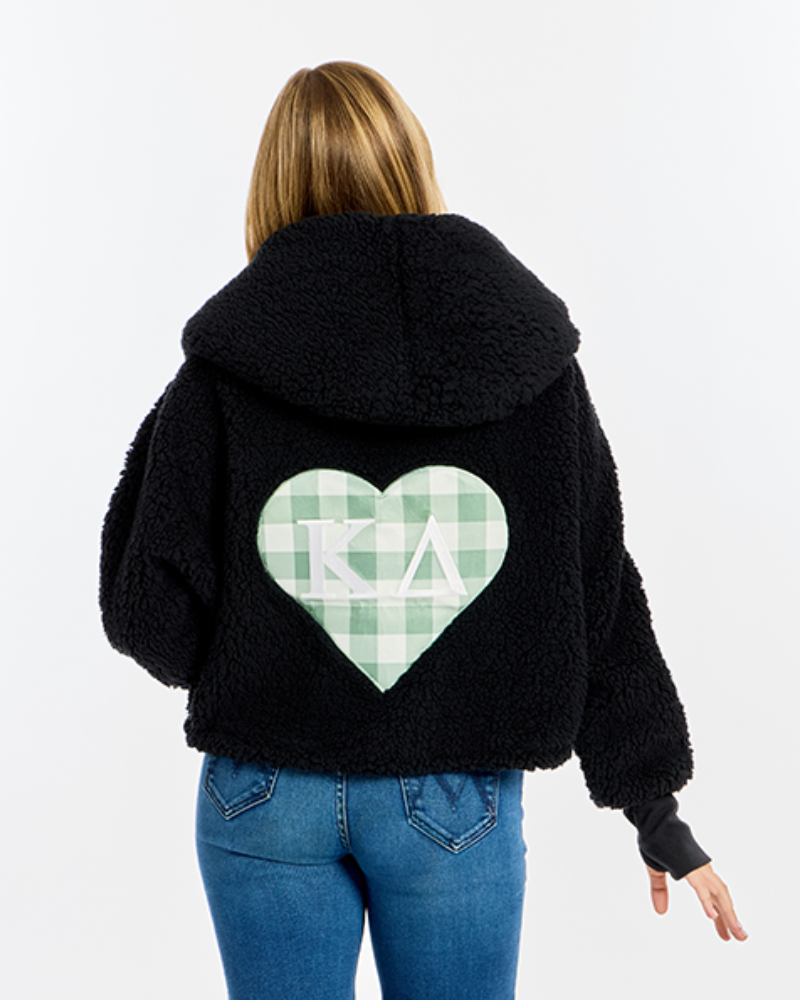 A woman viewed from behind wearing a black sherpa fleece hoodie with a large green and white gingham heart graphic on the back featuring the Greek letters 'Kappa Delta'. She is also wearing blue jeans against a plain white studio background.
