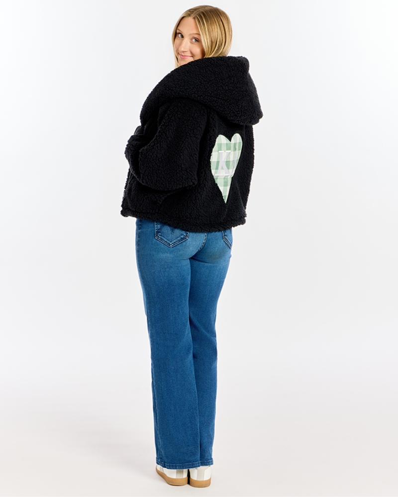 A woman wearing a black sherpa fleece hooded jacket with a green and white plaid heart patch on the back, paired with blue jeans, photographed from behind against a white studio background.