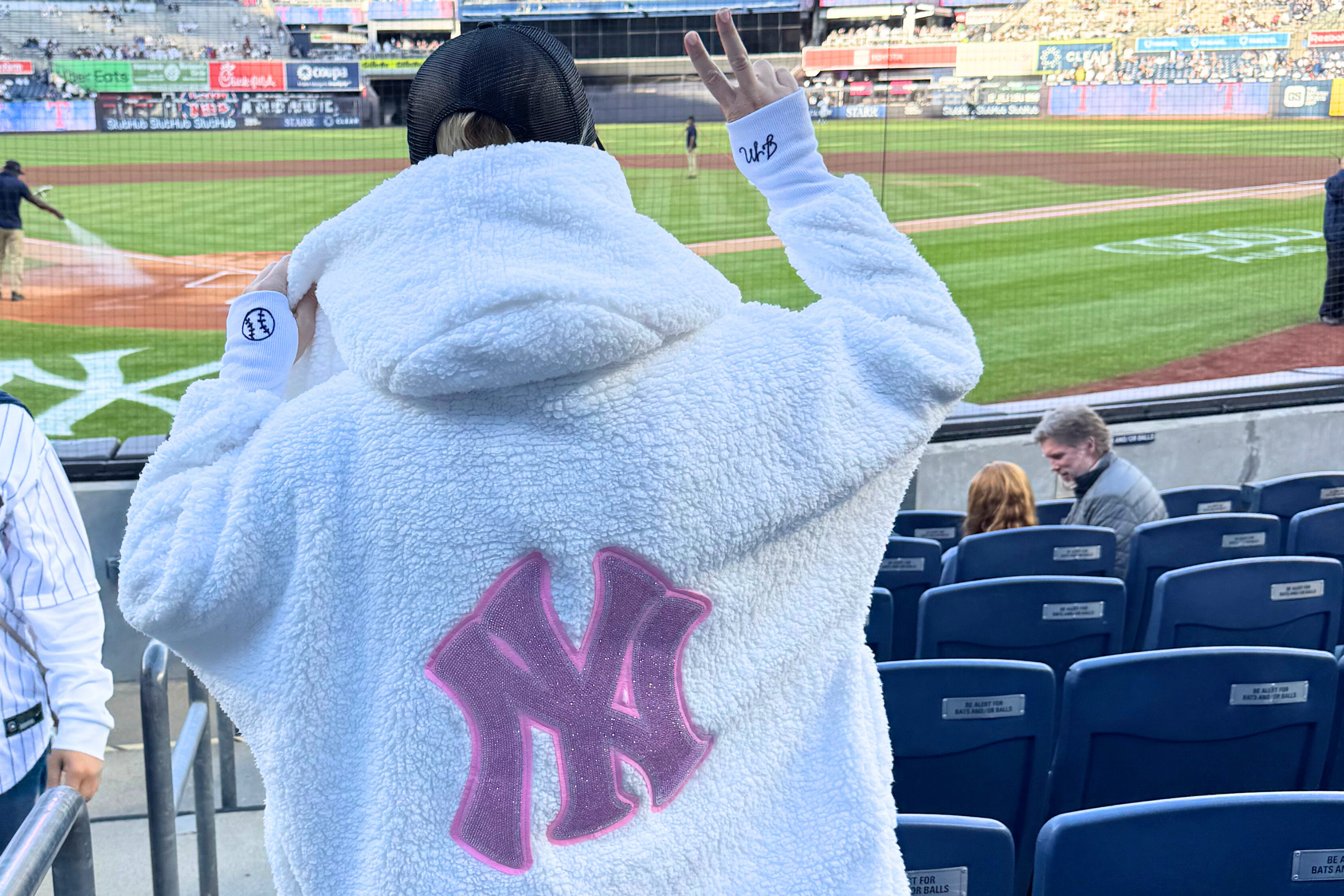 woman at baseball game standing in the stand wearing an oversized white sherpa lounger with a bedazzled new york yankees on back and holding up her hand with fingers in a peace sign