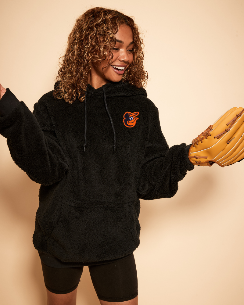Person wearing a black hoodie with a baltimore orioles logo and holding a baseball glove against a beige background