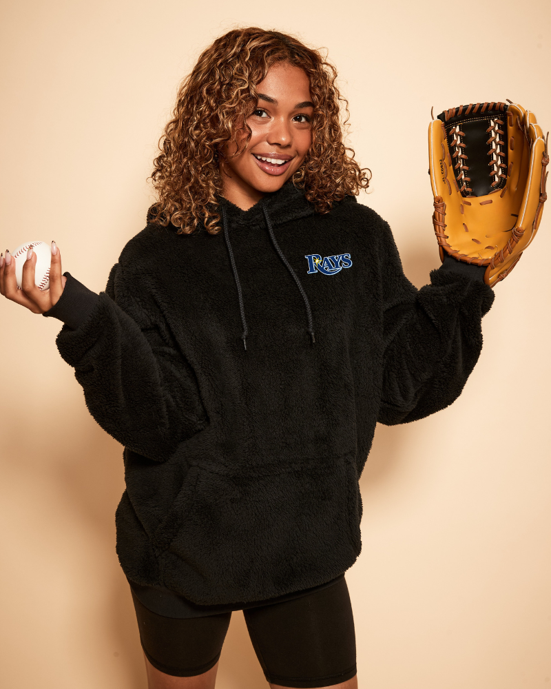 Person wearing a black hoodie with a tampa bay rays logo, holding a baseball and a glove against a beige background
