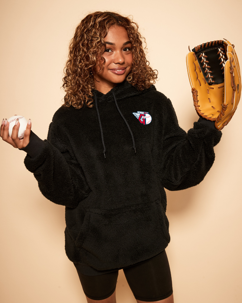 Person wearing a black hoodie with a cleveland guardians logo, holding a baseball and a glove on a beige background