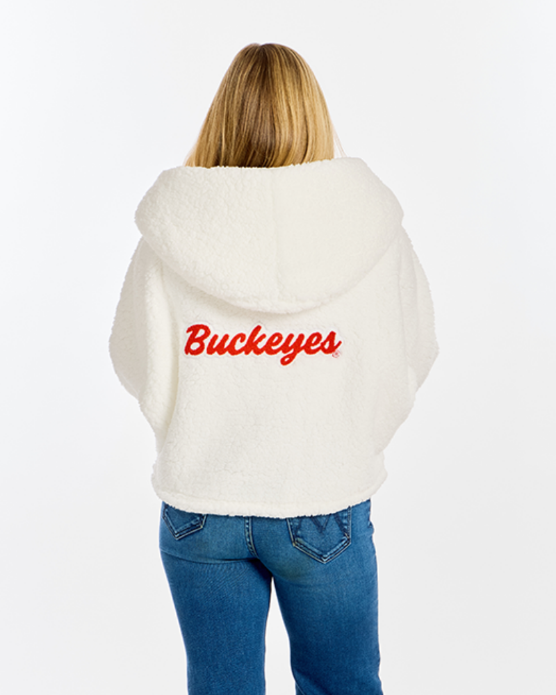 A woman viewed from behind wearing a cream-colored sherpa hoodie with 'Buckeyes' embroidered in red across the back, paired with blue jeans, against a plain white background.