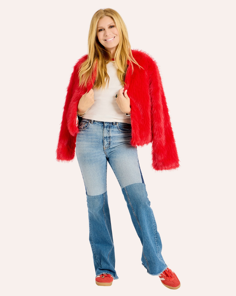 A woman modeling a bright red fuzzy faux-fur jacket over a white top and light blue patchwork jeans, standing against a plain light beige studio background.