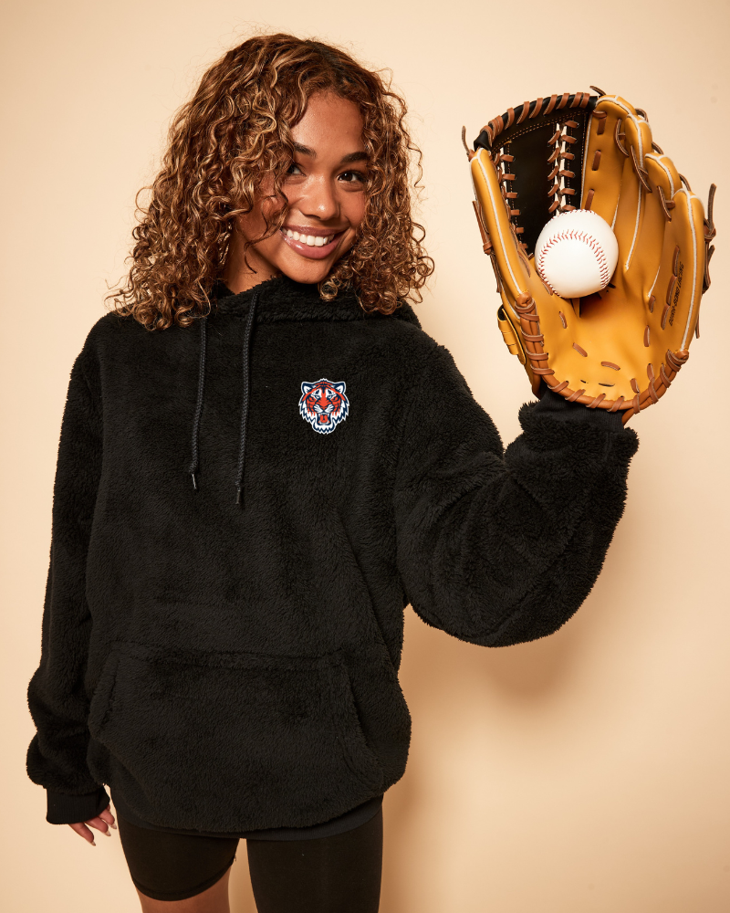 Person wearing a black hoodie with a detroit tigers logo, holding a baseball glove and ball against a beige background