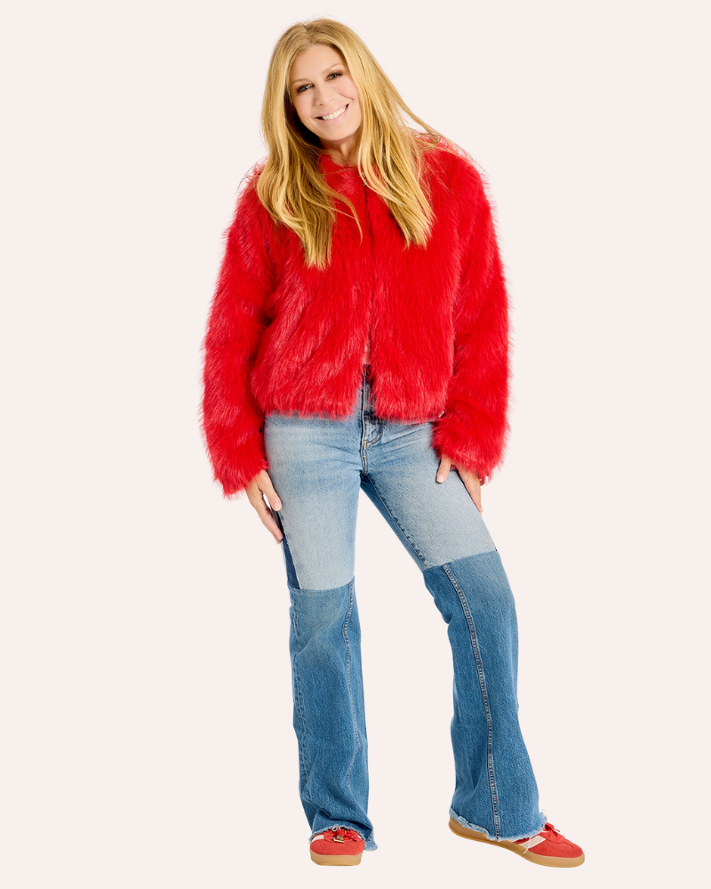 A woman modeling a bright red fuzzy faux-fur jacket, paired with blue patchwork jeans and red slip-on shoes, against a solid off-white background.